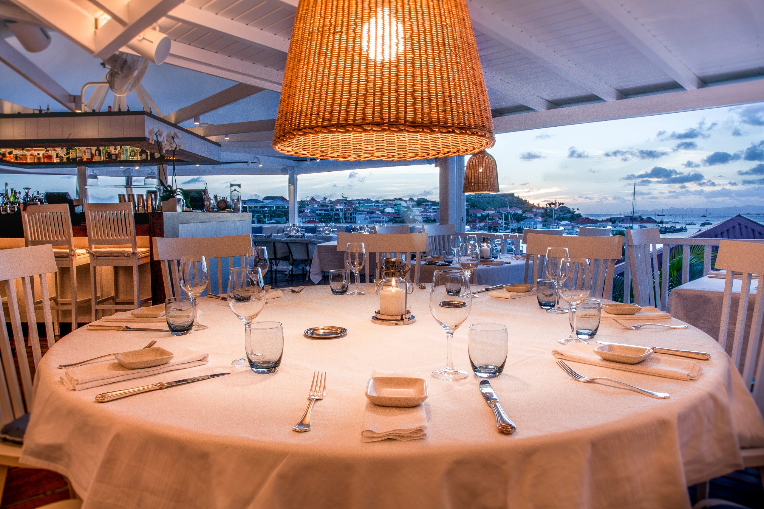A round table set for dinner with harbour and sunset visible through the rail